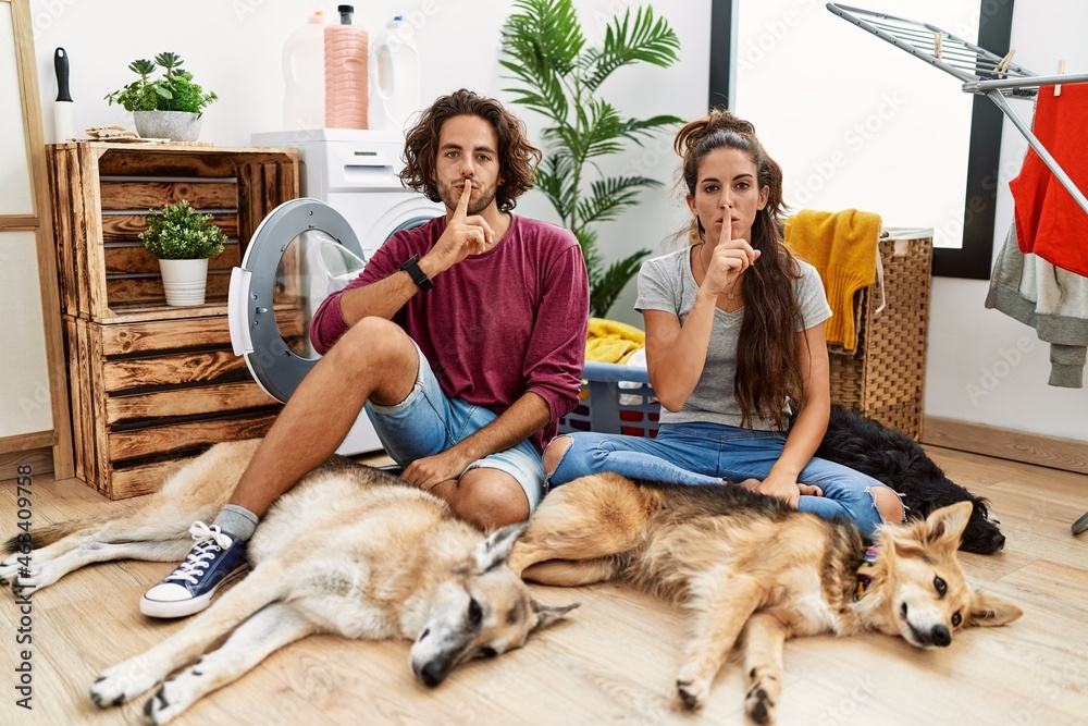 Young hispanic couple doing laundry with dogs asking to be quiet with finger on lips. silence and secret concept.