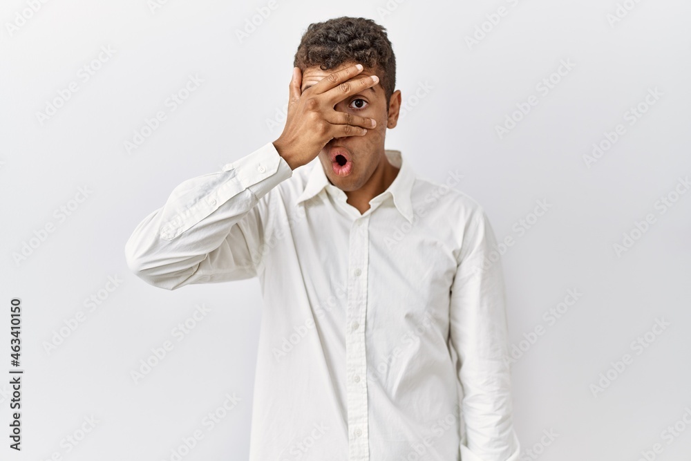 Young handsome hispanic man standing over isolated background peeking in shock covering face and eyes with hand, looking through fingers with embarrassed expression. Young handsome hispanic man standing over isolated background peeking in shock covering face and eyes with hand, looking through fingers with embarrassed expression.