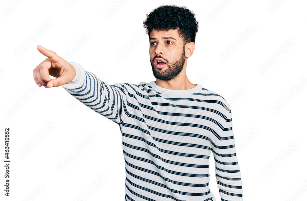 Young arab man with beard wearing casual striped sweater pointing with finger surprised ahead, open mouth amazed expression, something on the front Young arab man with beard wearing casual striped sweater pointing with finger surprised ahead, open mouth amazed expression, something on the front