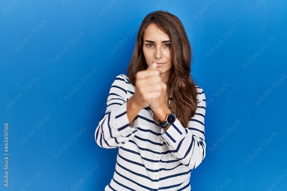 Young hispanic woman standing over blue isolated background ready to fight with fist defense gesture, angry and upset face, afraid of problem Young hispanic woman standing over blue isolated background ready to fight with fist defense gesture, angry and upset face, afraid of problem