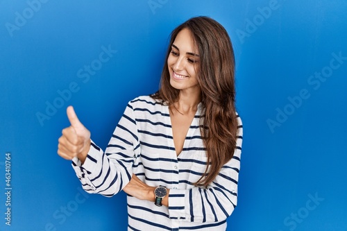Young hispanic woman standing over blue isolated background looking proud, smiling doing thumbs up gesture to the side