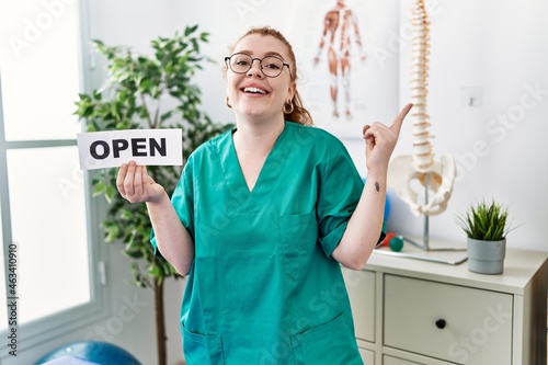 Young redhead physiotherapist woman working at pain recovery clinic holding open banner smiling happy pointing with hand and finger to the side