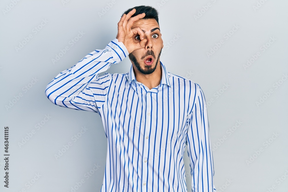 Young hispanic man with beard wearing casual striped shirt doing ok gesture shocked with surprised face, eye looking through fingers. unbelieving expression. Young hispanic man with beard wearing casual striped shirt doing ok gesture shocked with surprised face, eye looking through fingers. unbelieving expression.