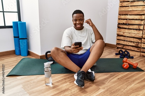 Young african man sitting on training mat at the gym using smartphone smiling doing phone gesture with hand and fingers like talking on the telephone. communicating concepts.