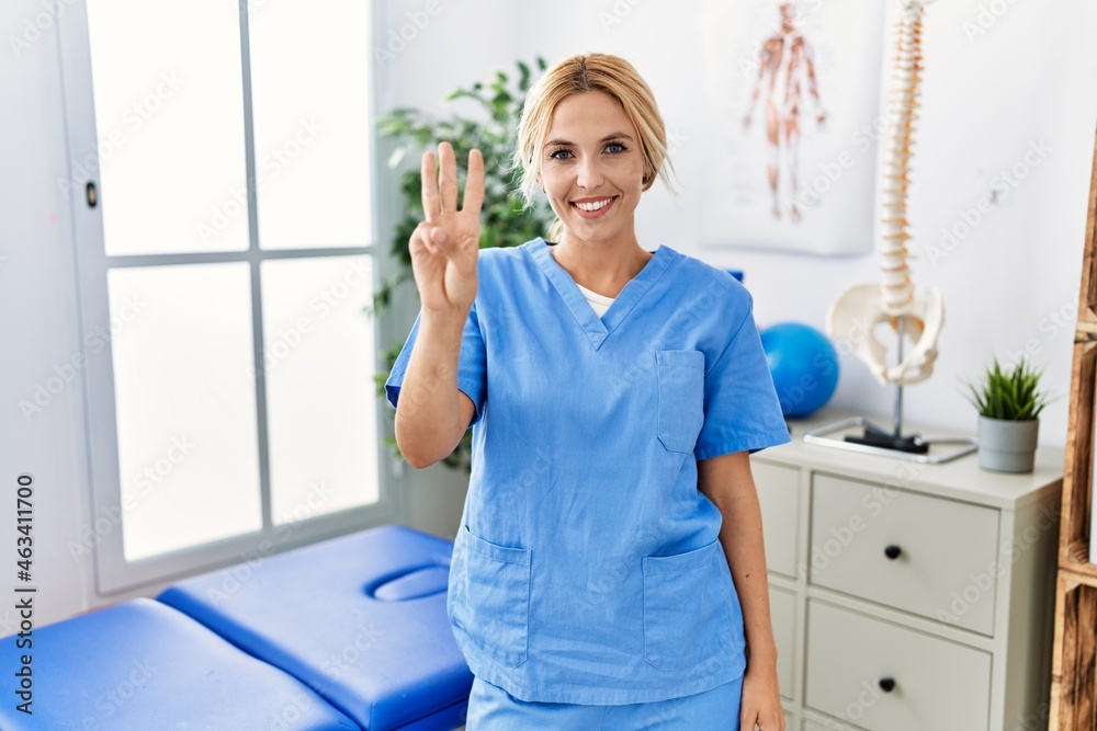 Beautiful blonde physiotherapist woman working at pain recovery clinic showing and pointing up with fingers number three while smiling confident and happy.