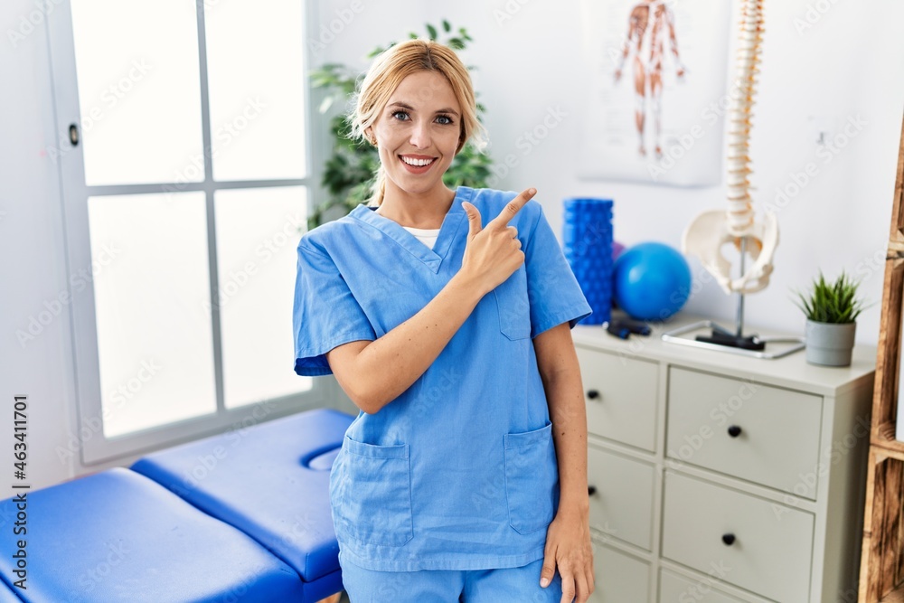 Beautiful blonde physiotherapist woman working at pain recovery clinic cheerful with a smile of face pointing with hand and finger up to the side with happy and natural expression on face Beautiful blonde physiotherapist woman working at pain recovery clinic cheerful with a smile of face pointing with hand and finger up to the side with happy and natural expression on face