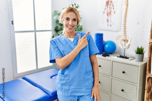 Beautiful blonde physiotherapist woman working at pain recovery clinic cheerful with a smile of face pointing with hand and finger up to the side with happy and natural expression on face