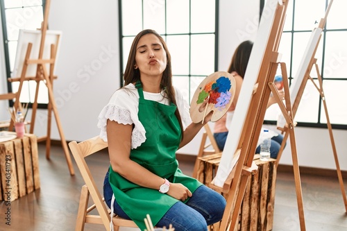 Young hispanic artist women painting on canvas at art studio looking at the camera blowing a kiss on air being lovely and sexy. love expression.