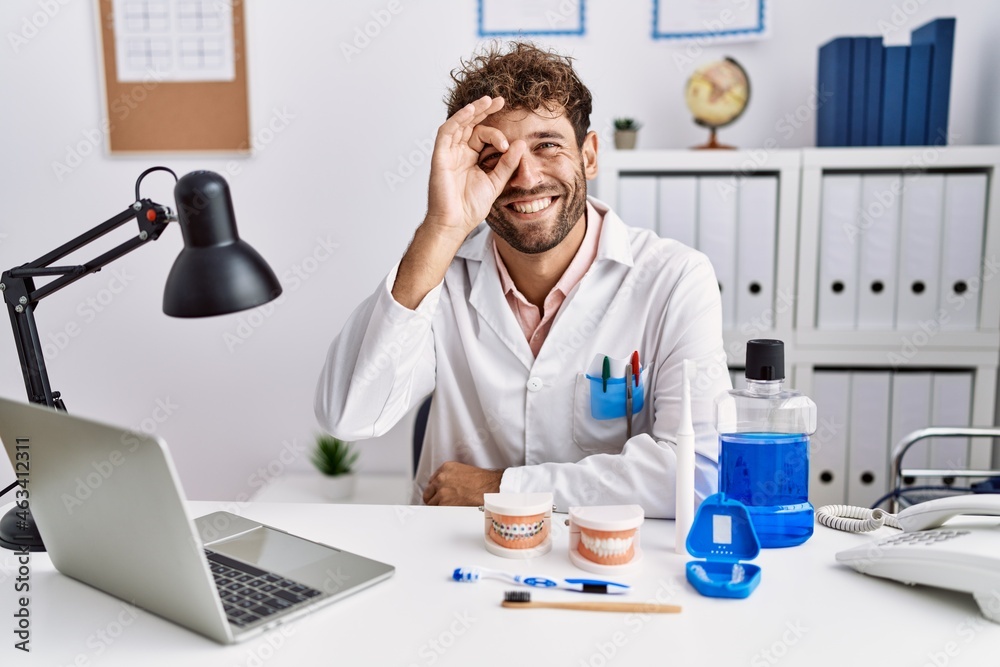 Young hispanic dentist man working at medical clinic doing ok gesture with hand smiling, eye looking through fingers with happy face. Young hispanic dentist man working at medical clinic doing ok gesture with hand smiling, eye looking through fingers with happy face.