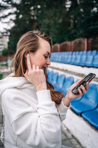 A beautiful woman in a tracksuit stands in a stadium wearing wireless headphones in one hand holds a case, in the other a phone with music for sports