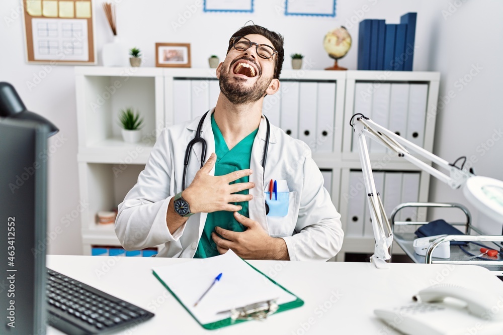 Young man with beard wearing doctor uniform and stethoscope at the clinic smiling and laughing hard out loud because funny crazy joke with hands on body. Young man with beard wearing doctor uniform and stethoscope at the clinic smiling and laughing hard out loud because funny crazy joke with hands on body.