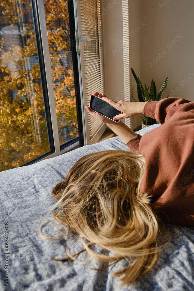 Naklejka premium Girl holds a smartphone in her hands in a comfortable bed and checks the social network account before going to bed or after waking up. Gadget addiction. Woman sending text message on mobile phone