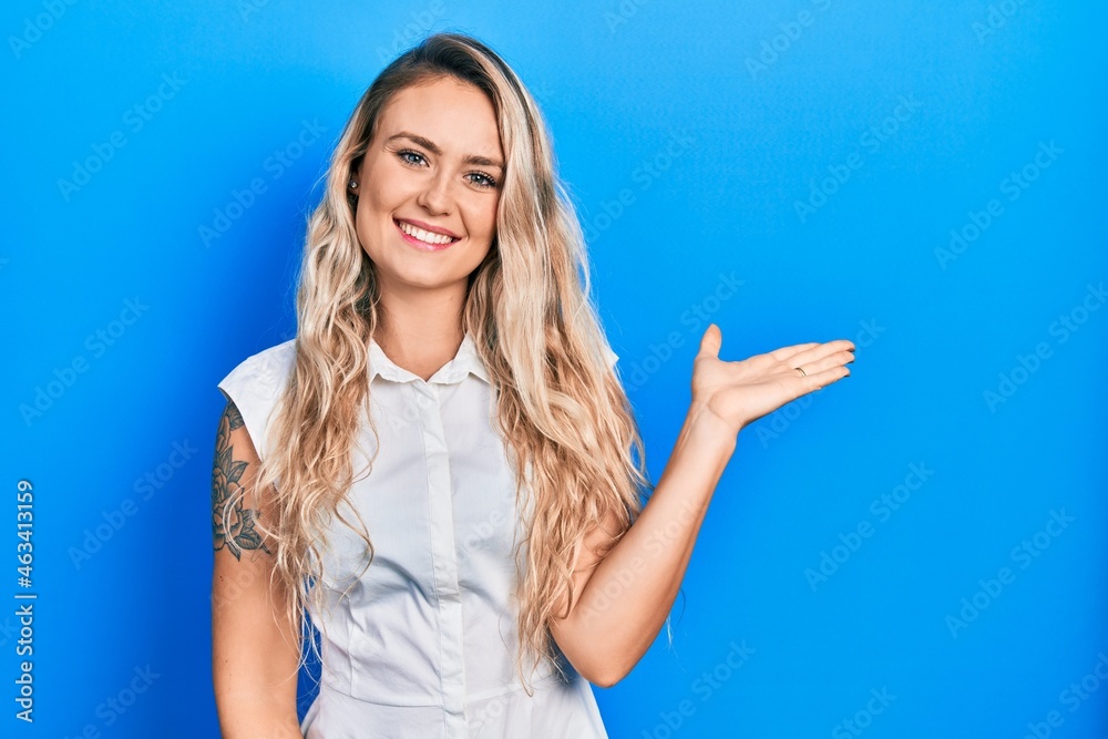 Fototapeta premium Beautiful young blonde woman wearing casual white shirt smiling cheerful presenting and pointing with palm of hand looking at the camera.