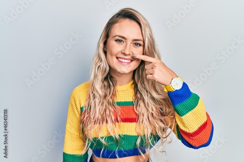 Beautiful young blonde woman wearing colored sweater pointing with hand finger to face and nose  smiling cheerful. beauty concept