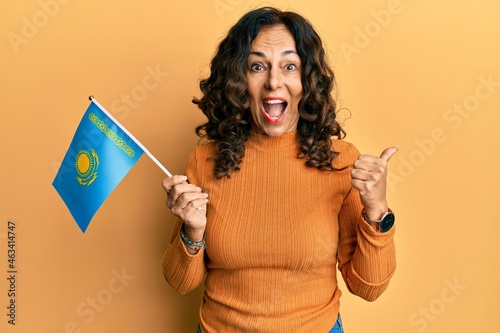 Middle age hispanic woman holding kazakhstan flag pointing thumb up to the side smiling happy with open mouth