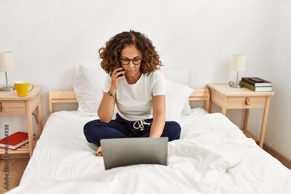 Middle age hispanic woman using laptop talking on the smartphone at bedroom Middle age hispanic woman using laptop talking on the smartphone at bedroom