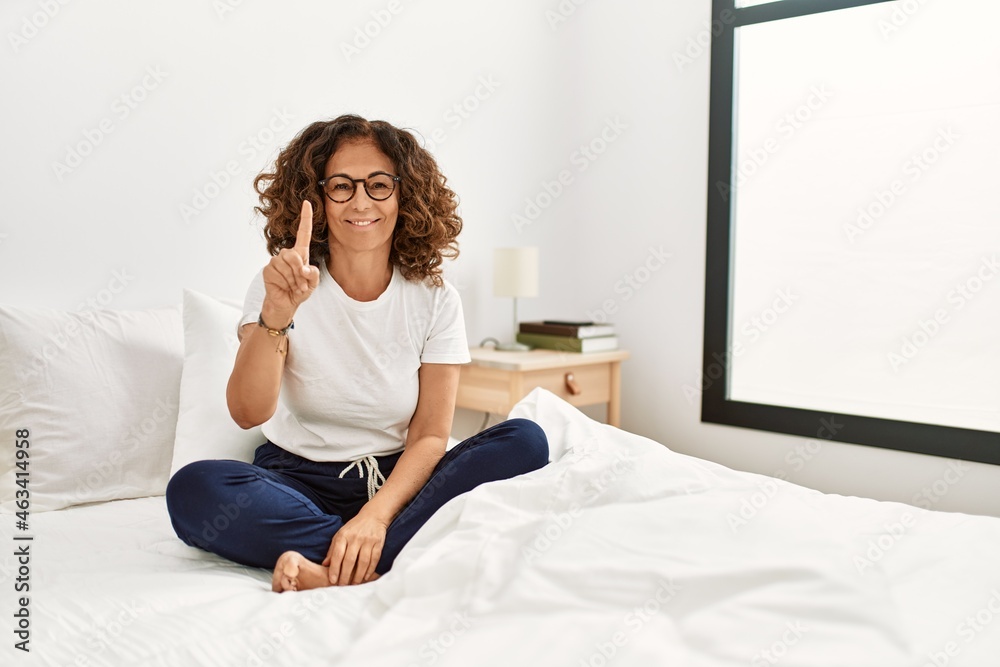 Middle age hispanic woman sitting on the bed at home showing and pointing up with finger number one while smiling confident and happy. Middle age hispanic woman sitting on the bed at home showing and pointing up with finger number one while smiling confident and happy.