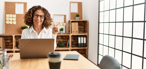 Middle age hispanic woman working at the office wearing glasses with a big smile on face, pointing with hand and finger to the side looking at the camera.