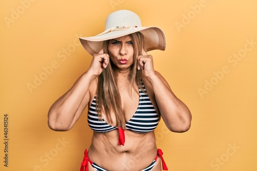 Middle age hispanic woman wearing bikini and summer hat covering ears with fingers with annoyed expression for the noise of loud music. deaf concept.