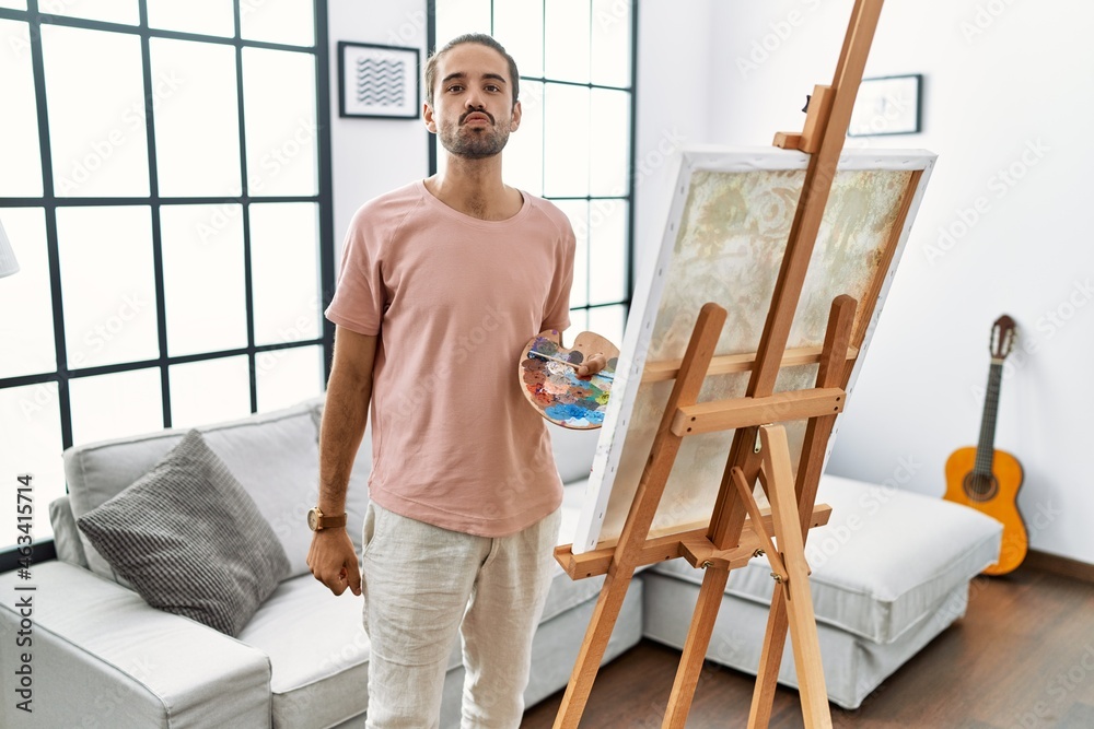 Young hispanic man with beard painting on canvas at home looking at the camera blowing a kiss on air being lovely and sexy. love expression.