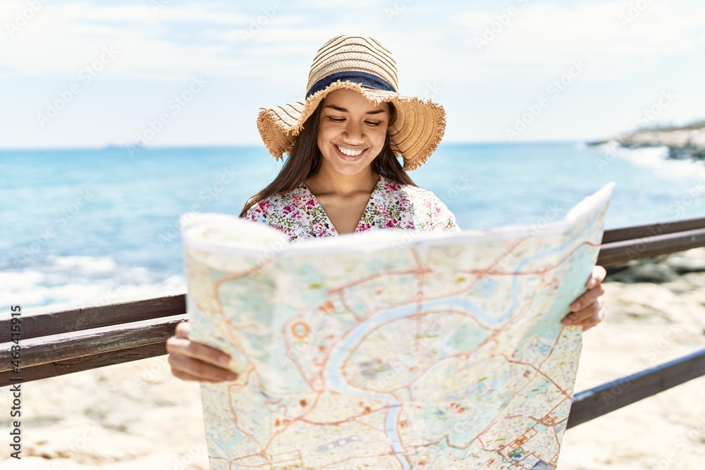 Young latin girl wearing summer hat holding city map at the beach.