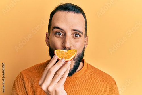 Young man with beard holding orange slice on mouth as funny smile scared and amazed with open mouth for surprise, disbelief face