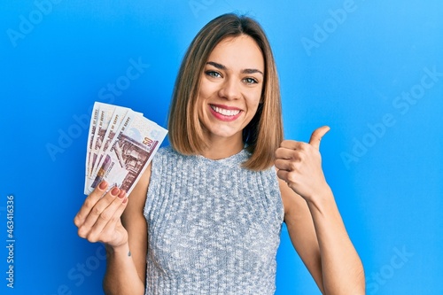 Young caucasian blonde woman holding egyptian pounds banknotes smiling happy and positive, thumb up doing excellent and approval sign