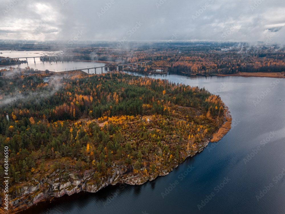 Aerial view on island with various trees with green and yellow foliage, rocky shore. On the background two bridges. Cloudy sky. Landscape of Scandinavian nature. Viborg, Russia.