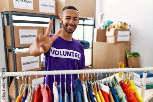 African american man wearing volunteer t shirt at donations stand smiling looking to the camera showing fingers doing victory sign. number two.
