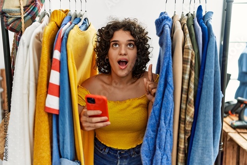 Young hispanic woman searching clothes on clothing rack using smartphone amazed and surprised looking up and pointing with fingers and raised arms.