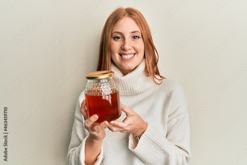 Young irish woman holding jar with honey looking positive and happy standing and smiling with a confident smile showing teeth Young irish woman holding jar with honey looking positive and happy standing and smiling with a confident smile showing teeth