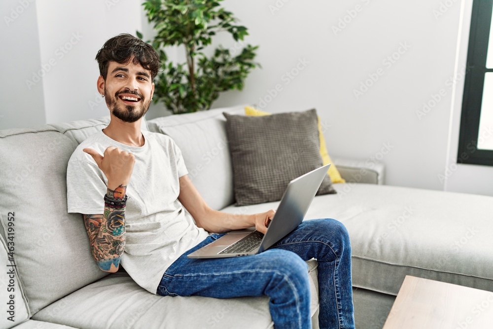 Hispanic man with beard sitting on the sofa smiling with happy face looking and pointing to the side with thumb up.