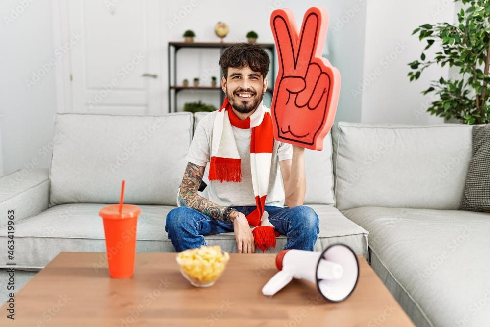 Naklejka premium Hispanic man with beard supporting football team at home sitting on the sofa looking positive and happy standing and smiling with a confident smile showing teeth