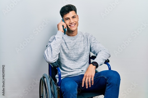 Young hispanic man sitting on wheelchair talking on the smartphone looking positive and happy standing and smiling with a confident smile showing teeth