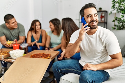 Group of young hispanic friends eating italian pizza sitting on the sofa. Man smiling happy and talking on the smartphone at home.