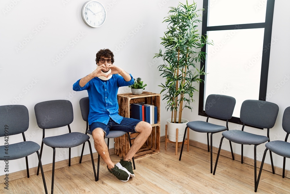 Fototapeta premium Young hispanic man sitting at doctor waiting room with neck injury smiling in love showing heart symbol and shape with hands. romantic concept.