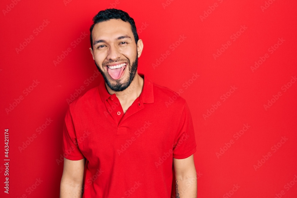 Hispanic man with beard wearing casual red t shirt sticking tongue out happy with funny expression. emotion concept. Hispanic man with beard wearing casual red t shirt sticking tongue out happy with funny expression. emotion concept.