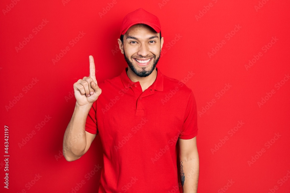 Hispanic man with beard wearing delivery uniform and cap showing and pointing up with finger number one while smiling confident and happy.