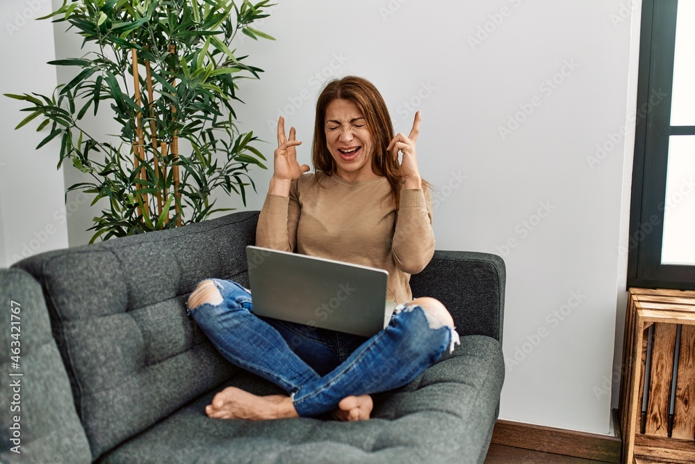 Senior middle east woman using laptop sitting on the sofa at home celebrating mad and crazy for success with arms raised and closed eyes screaming excited. winner concept