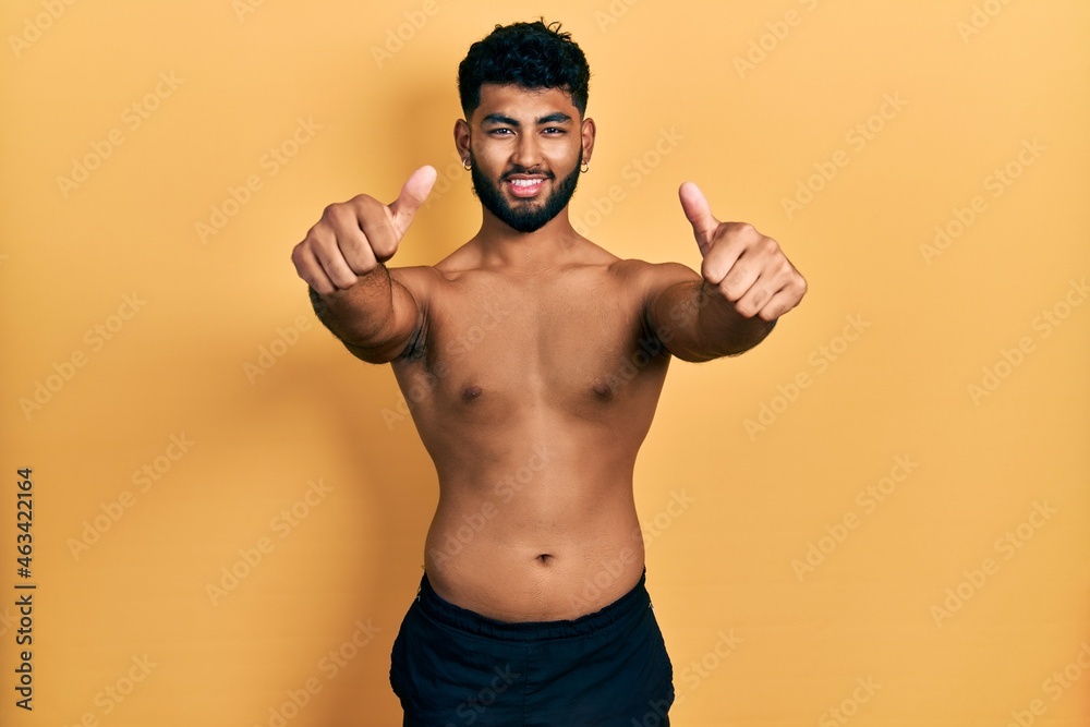 Arab man with beard wearing swimwear shirtless approving doing positive gesture with hand, thumbs up smiling and happy for success. winner gesture.