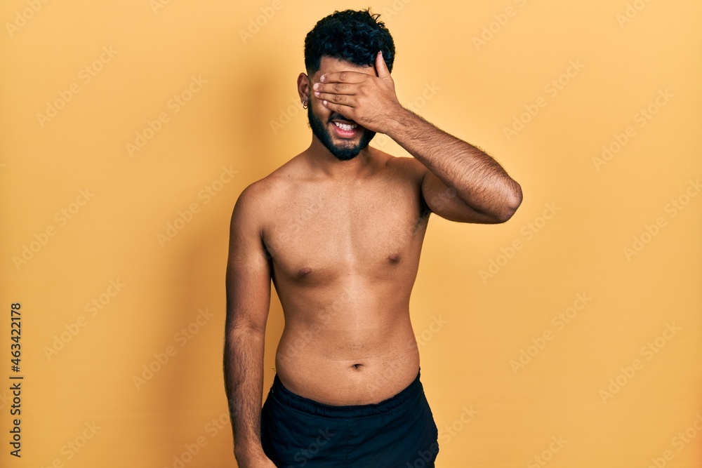 Arab man with beard wearing swimwear shirtless smiling and laughing with hand on face covering eyes for surprise. blind concept.