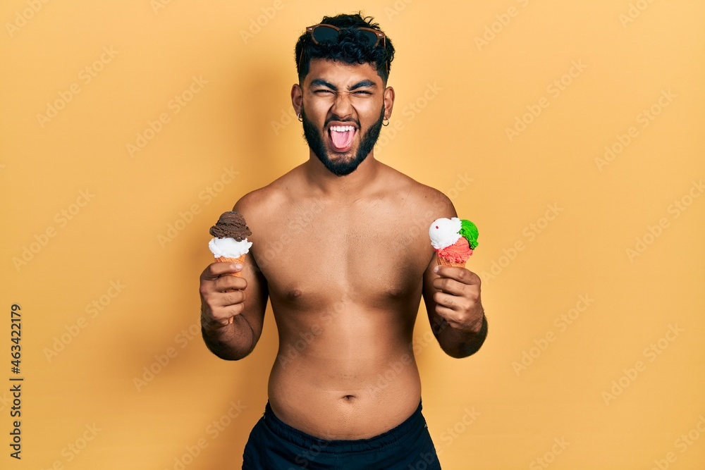 Arab man with beard wearing swimwear eating two ice cream cones sticking tongue out happy with funny expression. Arab man with beard wearing swimwear eating two ice cream cones sticking tongue out happy with funny expression.