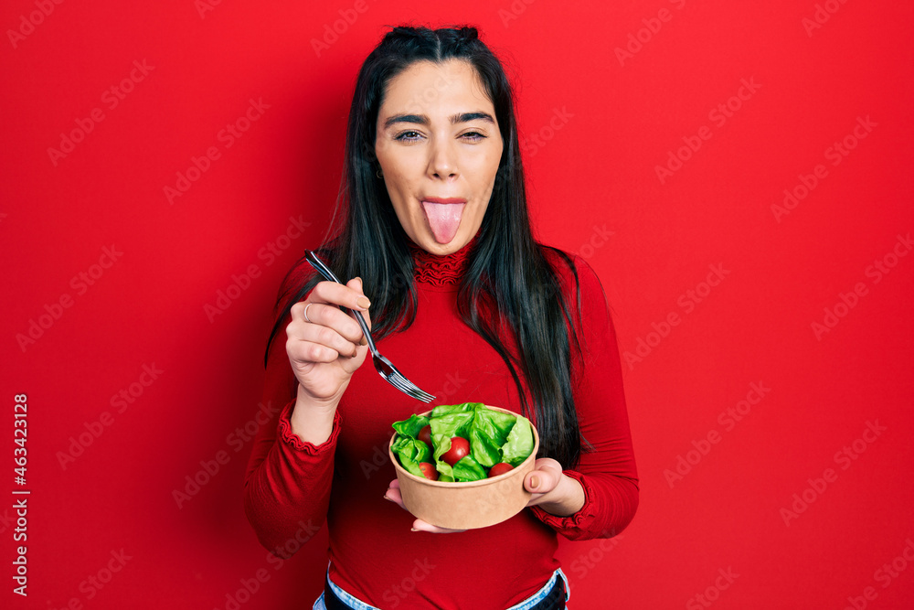Young hispanic girl eating salad sticking tongue out happy with funny expression.