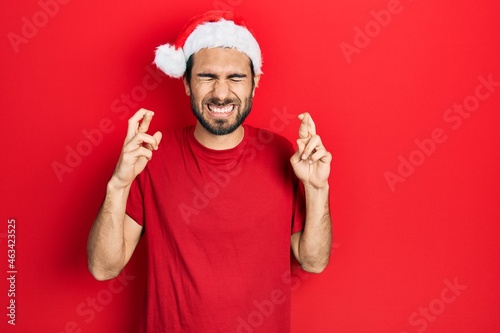Young hispanic man wearing christmas hat gesturing finger crossed smiling with hope and eyes closed. luck and superstitious concept. © Krakenimages.com