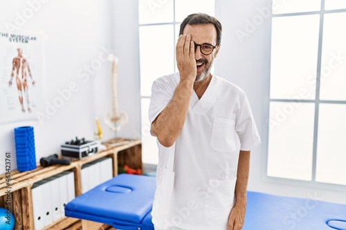 Middle age man with beard working at pain recovery clinic covering one eye with hand, confident smile on face and surprise emotion.