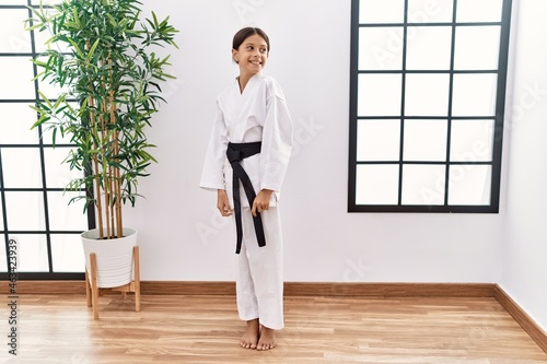 Young hispanic girl wearing karate kimono and black belt looking away to side with smile on face, natural expression. laughing confident.