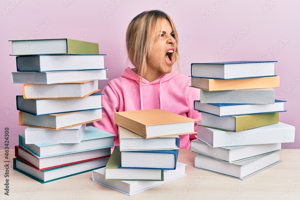 Young caucasian woman sitting on the table with books angry and mad screaming frustrated and furious, shouting with anger. rage and aggressive concept. Young caucasian woman sitting on the table with books angry and mad screaming frustrated and furious, shouting with anger. rage and aggressive concept.