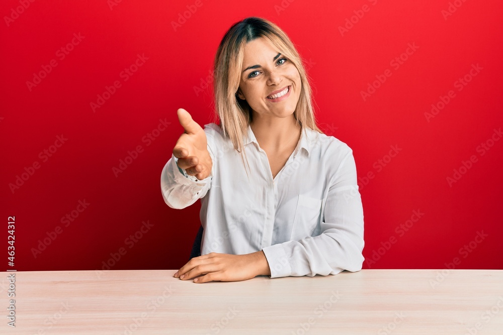 Young caucasian woman wearing casual clothes sitting on the table smiling friendly offering handshake as greeting and welcoming. successful business. Young caucasian woman wearing casual clothes sitting on the table smiling friendly offering handshake as greeting and welcoming. successful business.