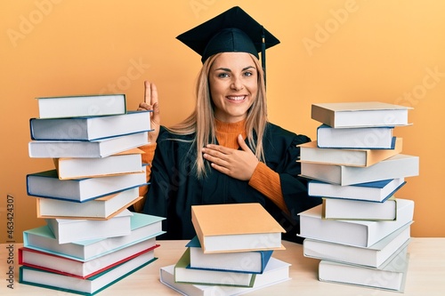 Young caucasian woman wearing graduation ceremony robe sitting on the table smiling swearing with hand on chest and fingers up, making a loyalty promise oath