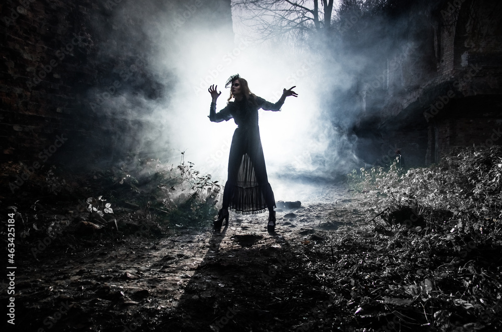 Naklejka premium A young witch dances and conjures on Halloween night in smoke and light at the festival of the dead.